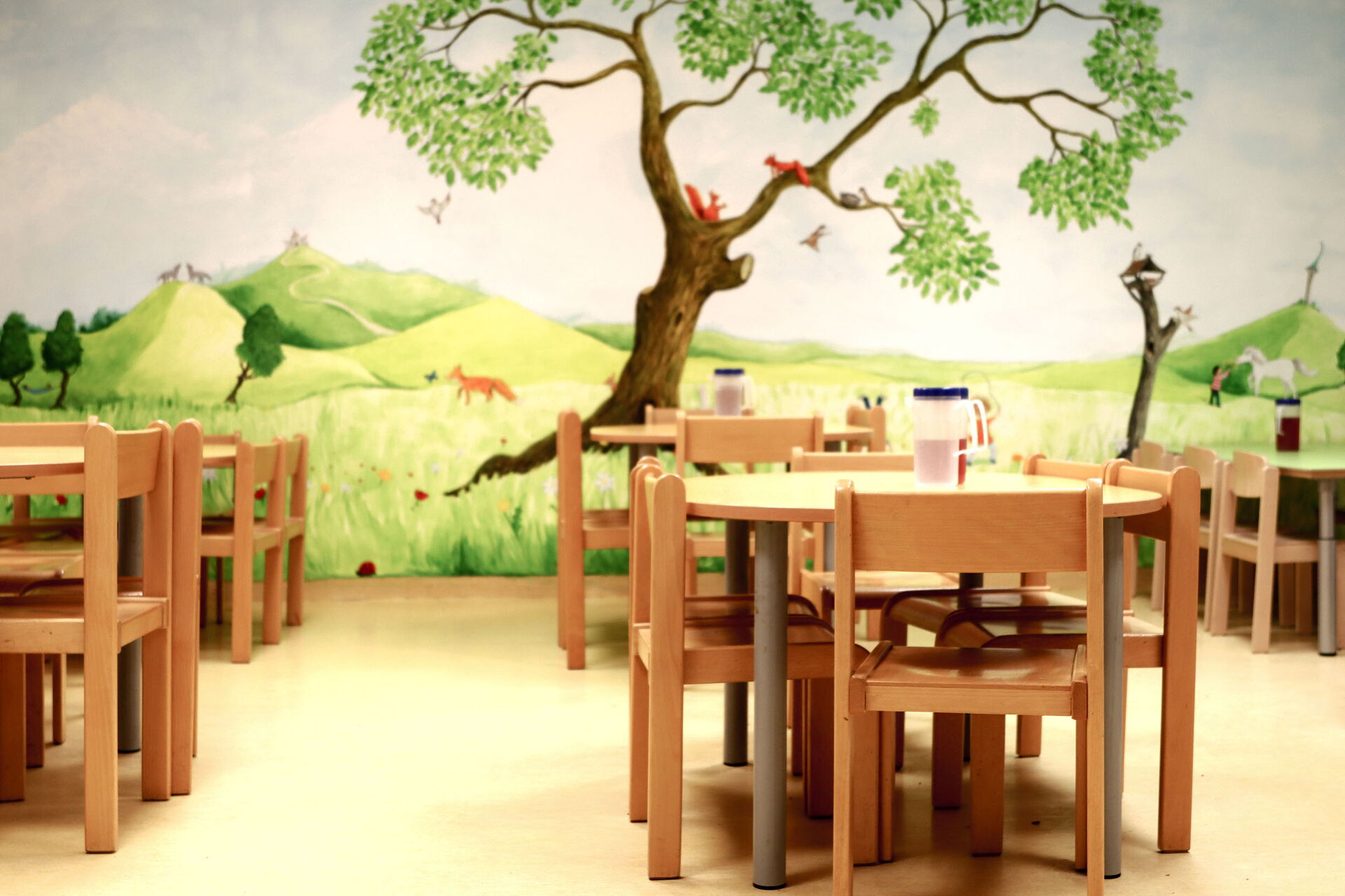 Speiseraum. Mehrere kleine Tische mit Stühlen. Großes Wandbild, mit Wiese, einem Baum in dem zwei Eichhörnchen sitzen.