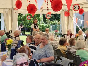 Festzelt, geschmückt mit Luftballons, Seidenpapiergirlande Rosette rot. Senioren sitzen an Tischen, trinken Kaffee und essen kuchen.