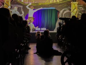 Theatersaal von hinten zur Bühne fotografiert. Im Gang zwischen den Zuschauenden sitzen Kinder auf dem Boden, es ist dunkel. Auf der Bühne sind 2 Schauspieler die tanzen.