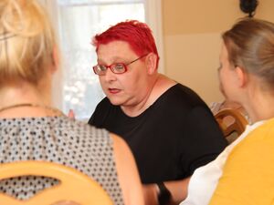 Im Zentrum des Fotos: Eine Frau mit kurzen roten Haaren, roter Brille und roten Lippenstift im schwarzen T-Shirt sitzt im inneren Kreis und spricht.