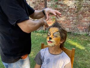 Kinderschminken. Kind sitzt auf einem Stuhl. Gesicht einer Raubkatze. Mann steht daneben und stylt noch die Haare passend.