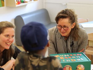 2 Frauen sitzen an einen Tisch mit einer grünen Box. Auf der Bux steht "Wunsch-Essen-Kinder"  Mit dem Rücken zu Kamera ein Kind mit blauem Cappy.