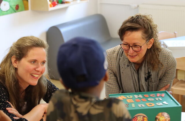 2 Frauen sitzen an einen Tisch mit einer grünen Box. Auf der Bux steht "Wunsch-Essen-Kinder"  Mit dem Rücken zu Kamera ein Kind mit blauem Cappy.