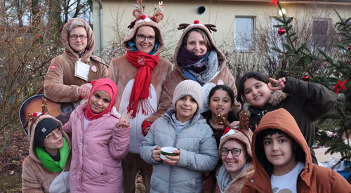 Vier weiße Frauen in Rentierkostümen. 3 stehen hinter südländischen Kindern in Winterkleidung. Eine der Frauen kniet zwischen den Kindern. Alle lächeln fröhlich in die Kamera. Eine Frau hat eine Gitarre umhängen.