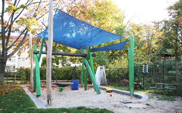 Spielplatz, großer Sandkasten mit Affenschaukel und einer blauen Plane als Überdachung.