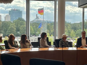 Im Bundestag, 5 Frauen, ein Mann an einem Tisch für großer Fensterfront. Draußen weht im Hintergrund die Deutschland Fahne.