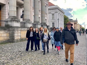 Gruppenbild. Frauen und ein Mann, der vorne rechts auf die Kamera zu kommt. Draußen Stadtszene.