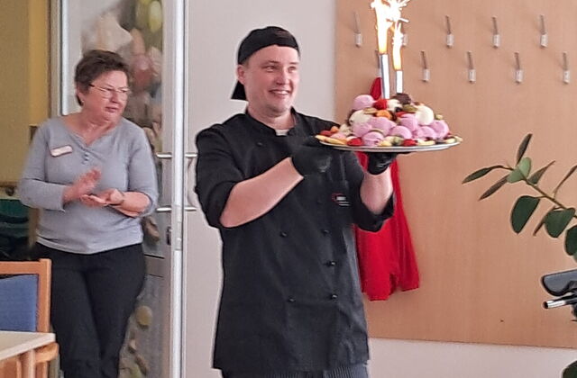 Küchenchef läuft bei den Frauen vorbei dabei hat auf einem Tablett bunte Eiskugeln gestapelt und zwei Leuchtfontänen verziehrt