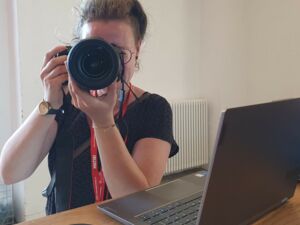 Eine Frau mit Kamera vor dem Gesicht, fotografiert den Fotografen. Vor ihr auf dem Tisch ein Laptop, und die Kameraklappe. Sie trägt ein schwarzes T-Shirt, eine Armbanduhr am rechten Handgelenk.