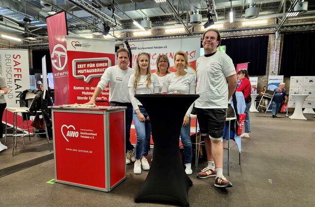 AWO Potsdam Messestand in einer Halle. 5 Mitarbeitende 3 Frauen eingerahmt von zwei Männern sie stehen hinter einem schwarzen Stehtisch. Messetheke mit AWO Logo Druck.