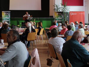 Voller Saal. Menschen sitzen an Tischen. Ganz vorn im Raum auf einem Podest Mann mit Gitarre und Notenständer.