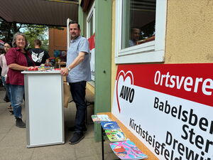 Ein Frau und ein Mann stehen sich gegenüber dazwischen ein Stehtisch mit Giveaways Sie stehen vor einem Gebäude und schauen beide in die Kamera.