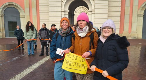 Frauen halten ein orangenes Band. 3 Frauen im Vordergrund mit orangenem Luftballon und einem Schild in den Händen. Text: Wo ist die Ministerin?