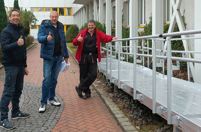 Drei Männer die den Damen nach oben zeigen stehen neben einer Rampe mit Geländer.