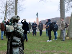 Draußen auf einer Wiese steht eine Gruppe im Kreis die undeutlich dargestellt ist. Im Vordergrund steht eine Skulptur.