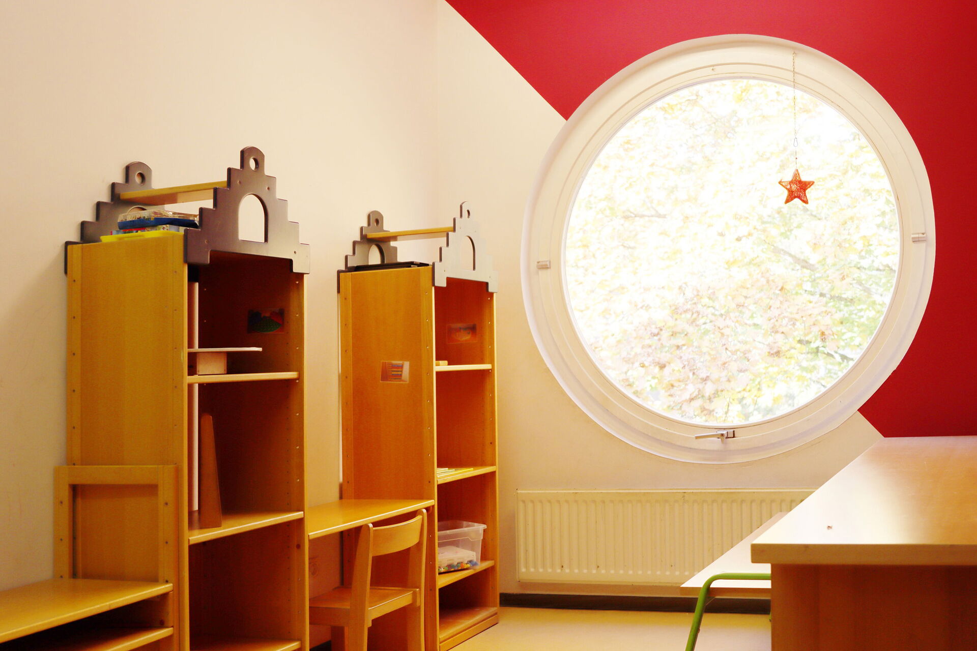 Zimmer mit großem Bullaugen-Fenster. Und Holzregal links an der Wand stehend.