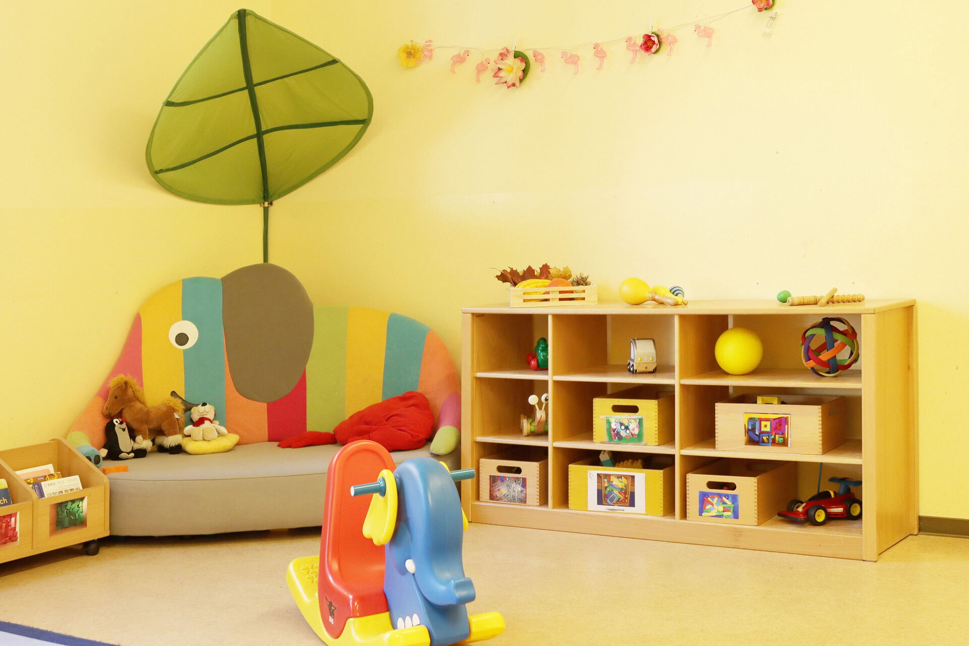 Kleiner bunter Schaukel-Elefant. Ein Elefanten Sitzecke und ein Regal mit Spielzeug steht an einer gelben Wand.