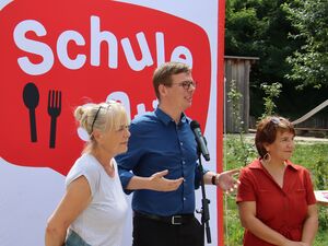 Draußen, Großes Plakat von Schule satt! 3 Personen stehen davor. Ein Mikrofon steht vor einem Redner. Junger Mann mit Brille, kurzen braunen Haaren und blauen Hemd. Links und rechts stehen zwei Frauen.