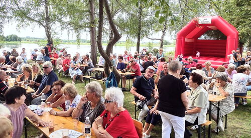 Viele Menschen an Biertischgarnituren. Essen und Getränke auf den Tischen. Im Hintergrund eine rote Hüpfburg.