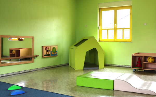 Spielzimmer. Raum mit grünen Wänden. An der Wans ein Spiegel und zwei Bilder, ein Pferd und ein Frosch. In der Ecke steht ein kleines grünes Häuschen mit einer Matte darin. Ein Fenster mit gelben Rahmen durch das die Sonne scheint.