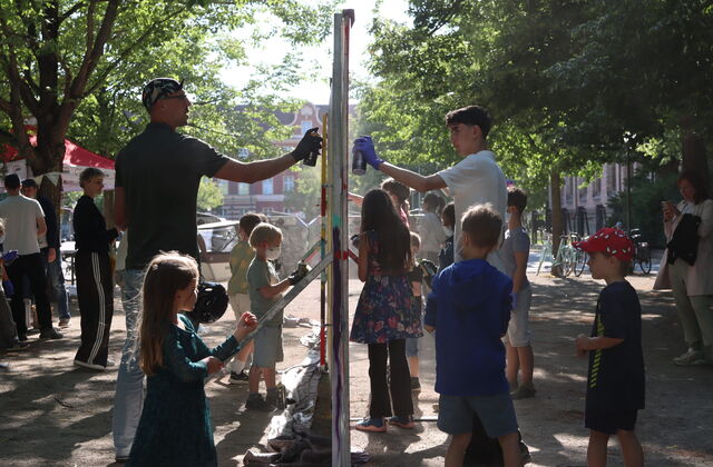 Ein Aufstellwand die von beiden Seiten mit Spraydosen besprüht wird. Kinder machen mit und schauen zu.