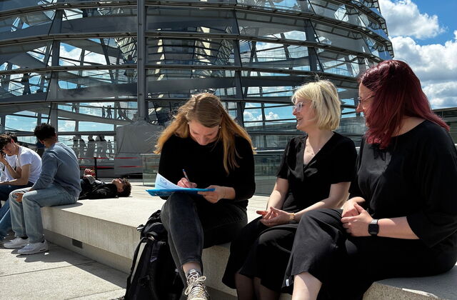 In der Sonne auf dem Doch vor der großen Glaskuppel. 3 Frauen sitzen dort und eine fülllt die Unterschriften Liste aus.