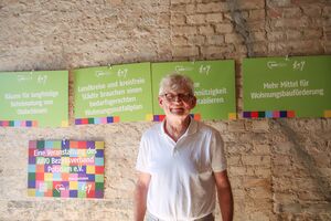 Portrait. Älterer Mann mit grauen, kurzen Haaren, mit Brille, weißen kurzärmligen Poloshirt. Er steht vor einer Steinwand. Am die Wand sind Schilder mit Forderungen.