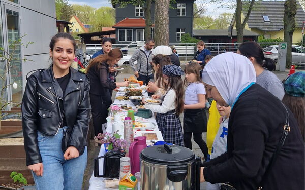 Draußen, ein Büfett, langer Tisch mit Getränken und Speisen. Kinder und Frauen holen sich was zu essen.
