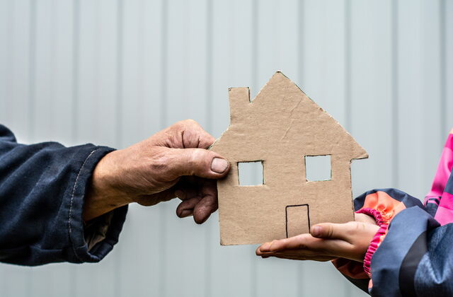 Zwei gegenüberstehende Personen halten jeweils mit einer Hand gemeinsam ein kleines Haus aus Pappe, im Hintergrund eine helle Wand.