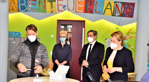 Ein farbenfroher Raum, grüne Wand. Vier Personen mit FFP 2 und OP Masken stehen um einen runden Stehtisch. Darauf eine Packung mit Corona Tests. Darüber bunte Buchstaben auf farbigen Papier: ABENTEUERLAND