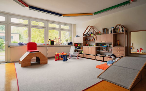 Spielzimmer. Regal mit Spielsachen an der Wand. Ein hellgrauer Teppich liegt im Zentrum des Raums. Puppenbettchen und Puppen.