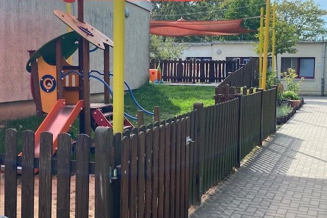 Gebäude. Draußen, ein kleines Klettergerüst mit Treppe uns Rutsche für Kleinkinder. Darüber ist ein organgenes Sonnensegel gespannt. Ein Holzzaun geht um das Kita-Gelände.