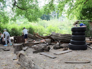 Baustelle im Garten. Reifen sind aufgestapelt und Holzstücke, Holzstämme liegen rum. 4 Kinder spielen dort.