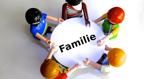 Fünf bunte Spielfiguren sitzen um einen runden weißen Tisch mit dem Schriftzug „Familie“ in der Mitte.
