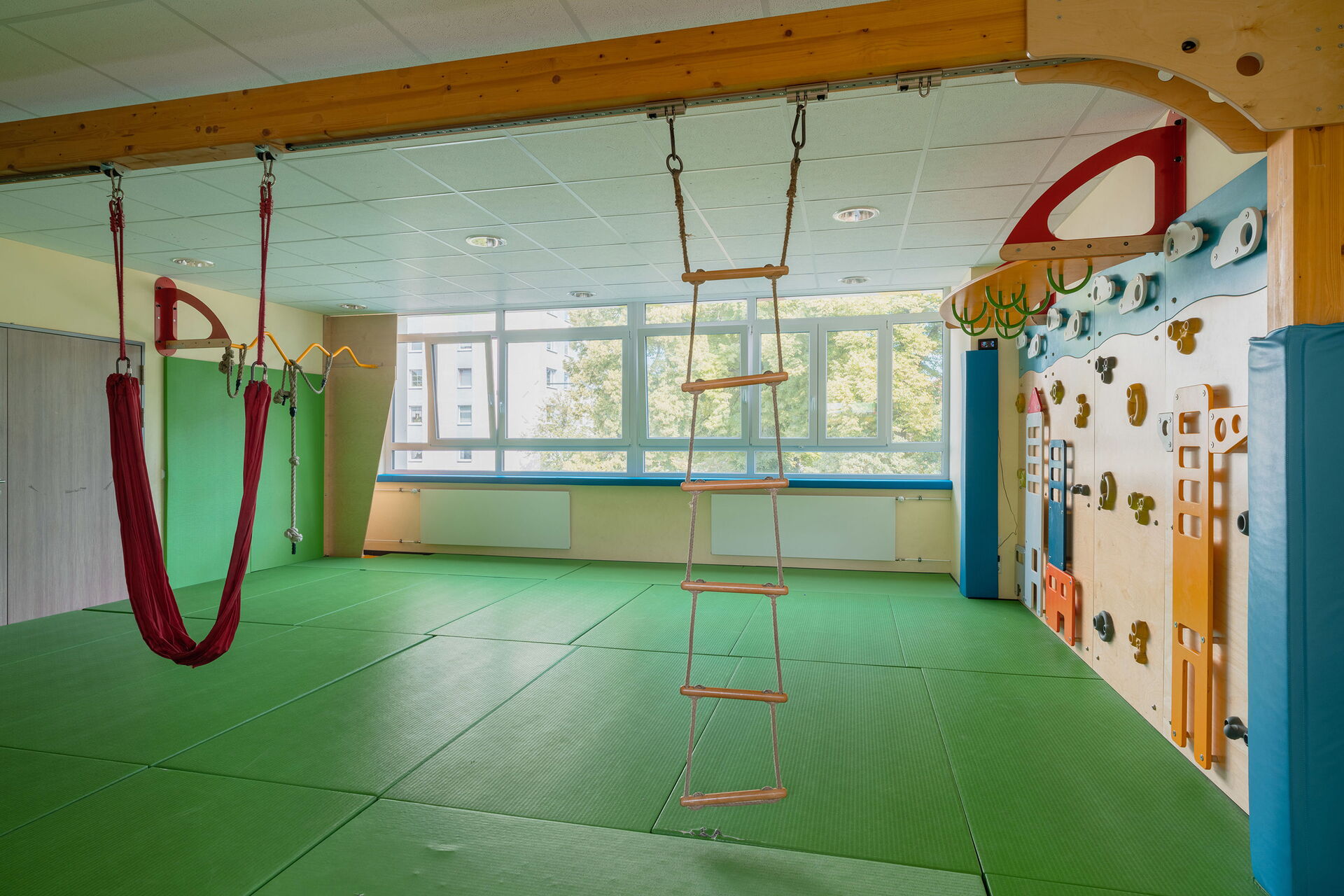 Sportraum. Hängeleiter an einem Holzbalken. Der Fußboden ist mit grünen Matten ausgelegt, An der Wand viel Griffe, Reifen und Tritte zum Klettern.