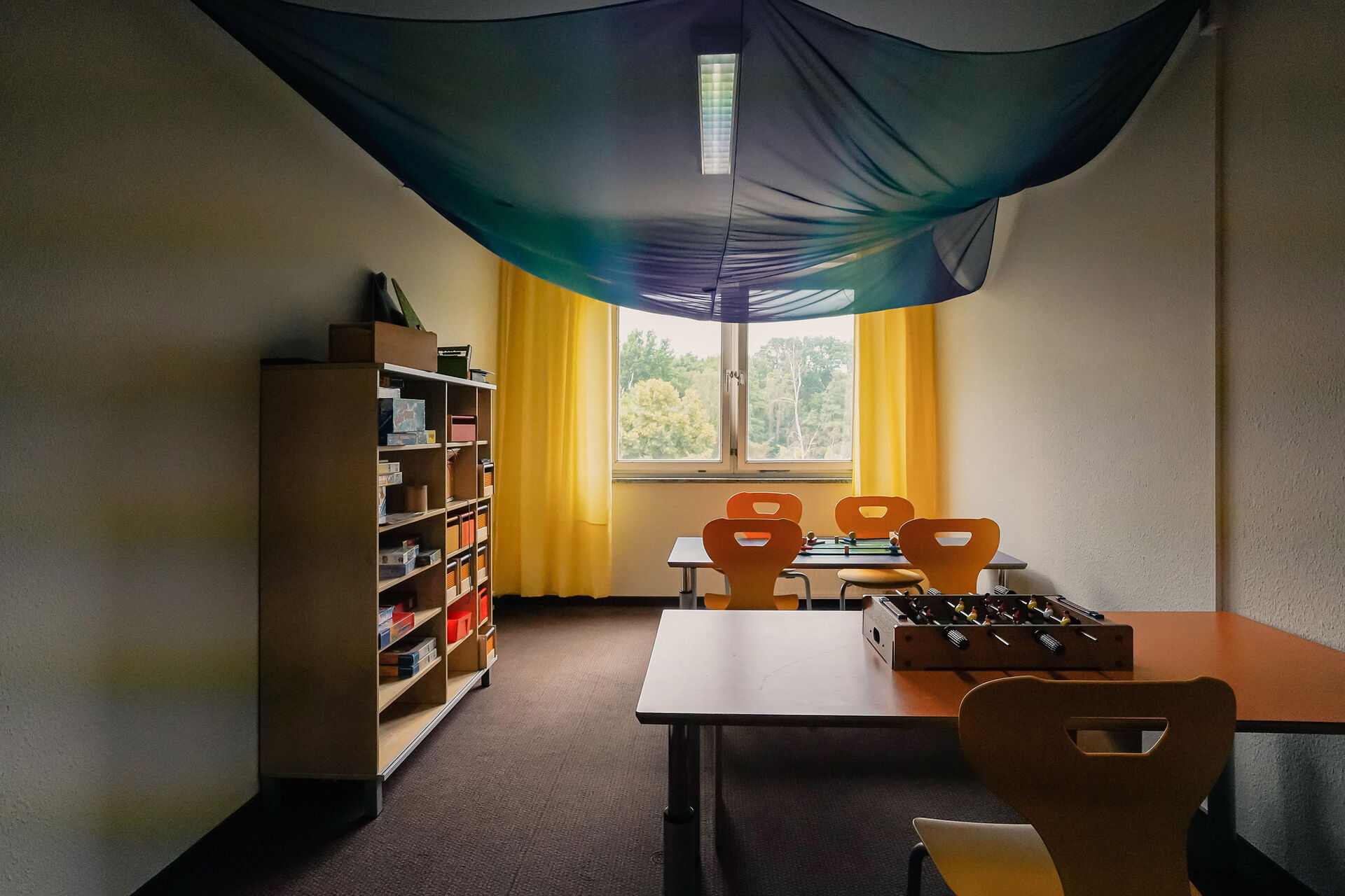 Spielzimmer, 2 Tische mit Stühlen an der Wand. Auf einem Tisch ein kleiner Tischkicker, an der linken Wand ein Regal mit Spielen.