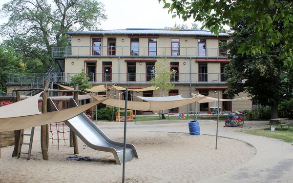 Spielplatz mit großen Sandkasten, Klettergerüst mit Rutsche. Im Hintergrund das dreistöckige Kitagebäude.