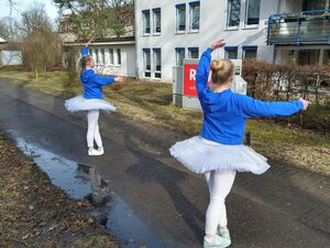 Draußen vor einem Gebäude. Pützen, ein Weg. Zwei weiße Ballett Tänzerinnen. Sie haben blonde lange haare, blaue Pullover, weißes Tutu, weiße Leggings und Turnschuhe an und tanzen.