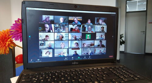 Eine Videokonferenz. Ein aufgeklappter Laptop. Auf dem Bildschirm viele Kacheln mit Menschen.