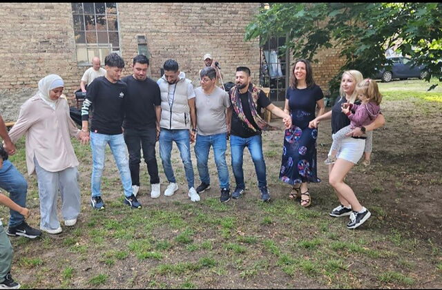 Draußen auf einer Wiese an einen gemauerten Gebäude. 10 Menschen die sich an den Händen halten und tanzen. Menschen mit unterschiedlichen Hautfarben. Eine Frau mit Kopftuch. Männer und zwei weiße Frauen, eine mit einem kleinen Mädchen auf dem Arm.