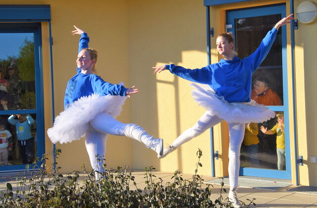 Zwei Ballerina in blau-weißen Kostümen. Blauer Pullover, weiße Strumpfhose tanzen draußen bei schönem Wetter. Kleinkinder gucken von drinnen durch verglaste Türen zu.