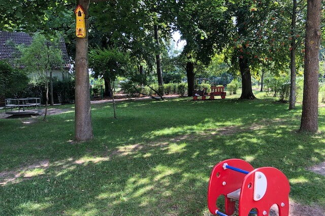 Weitläufiger Spielplatz, Rasen schattig durch einige Bäume. Vorne ein kleiner roter Elefant zum raufsetzen und reiten für kleine Kinder.