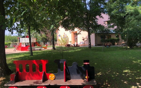 Eine Sitzbank für Kinder rot-schwarz. Sieht aus wie eine Eisennbahn. Die Lock ist schwarz. In Hintergrund Spielplatz, Sandkasten und ein Haus.