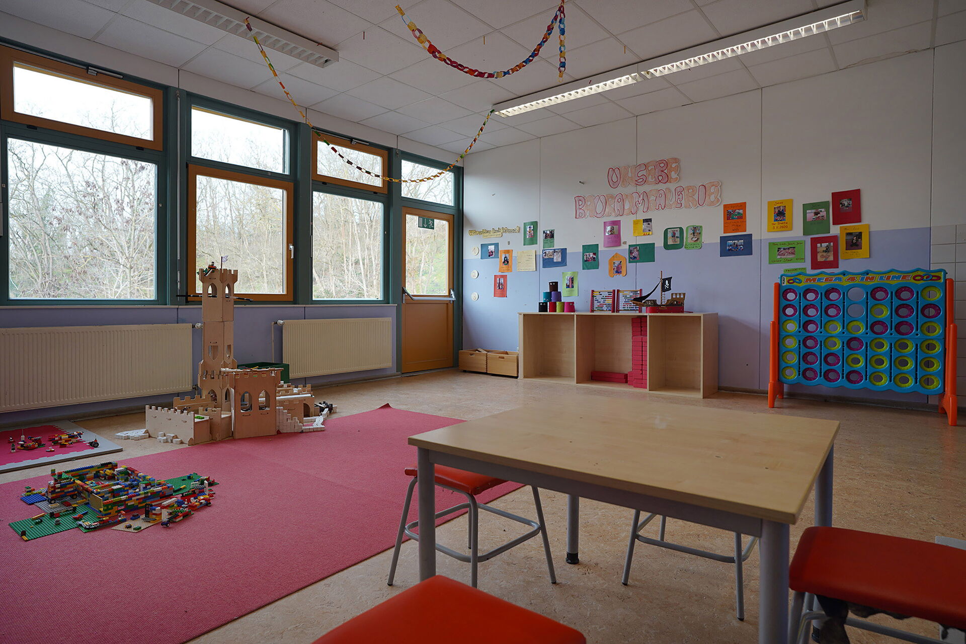 Spielzimmer. Großer Raum. links ist eine Burg aufgebaut. Ein Tisch mit vier Hocker. An der hinteren Wand ein Regal und an der Wand hängen Bilder.