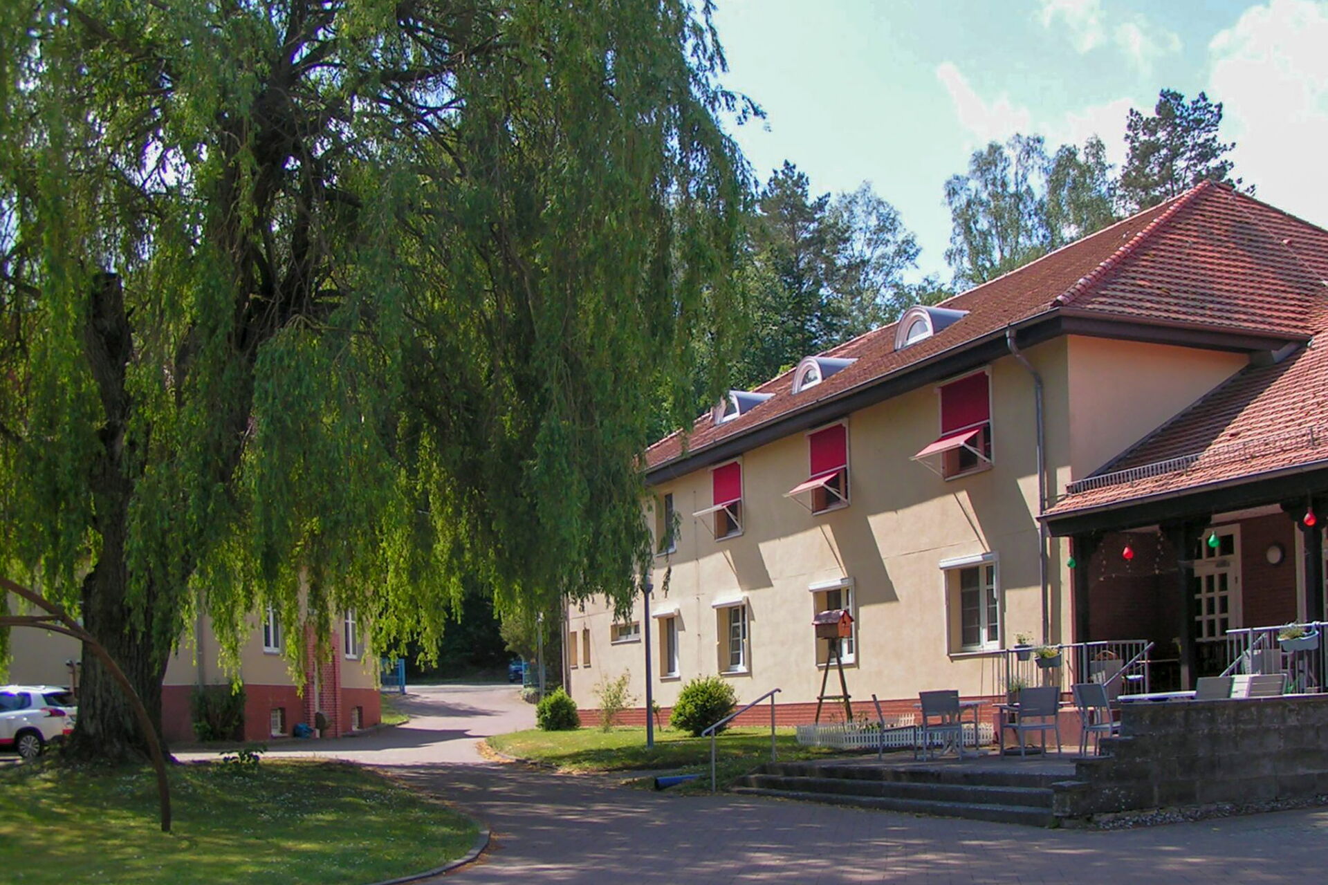 Große Weide links im Bild. Rechts ein Wohnhaus mit einer kleinen Terrasse auf der Stühle stehen.