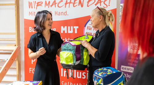 Zwei Frauen sehr fröhlich halten einen Schulranzen zwischen sich. Auf einem Tisch vor ihnen liegen mehrere bunte Federmappen für Kinder. Ein blauer Kanzen steh auf auf dem Tisch.