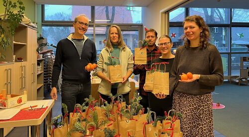 Im Raum. Runder Tisch mit vielen Nikolaus-Papier-Tüten, jede mit Weihnachtsaufkleber und Tannenzweigen verziert. Dahinter stehen zwei Männer und drei Frauen die lächeln und Tüten oder Mandarinen in den Händen halten.