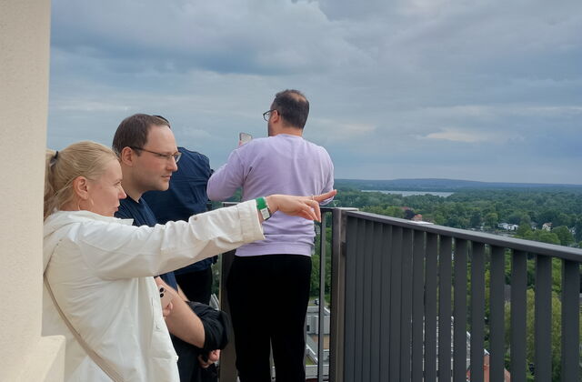 Menschen schauen von einer Aussichtsturm über Potsdam. Blonde Frau mit weißer Jacke streckt Arm aus und zeigt mit Finger nach rechts. Neben ihr ein Mann der in die gezeigte Richtung schaut.