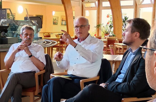 In einer Kantine/Cafeteria. 3 Männer sitzen im Halbkreis. Mann mit Brille und weißem langärmligen Hemd spricht. Es benutzt auch die Hände um etwas zu verdeutlichen.