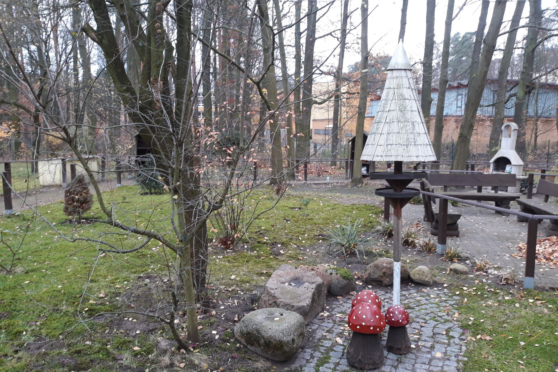 Kleine Parkanlage. Kahle Bäume und Sträucher, im Vordergrund 3 Deko Fliegenpilze, rechts 3 Bänke.